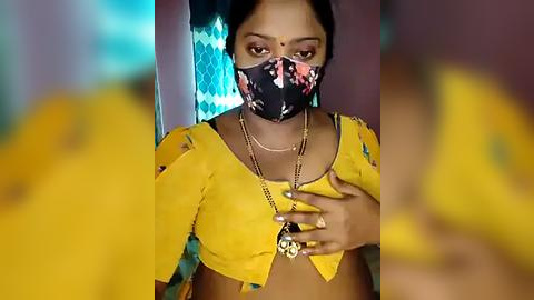 Media: Video of a young woman with medium skin tone, wearing a yellow blouse with blue floral patterns, black face mask, and gold necklace. Background features a blurred, multicolored pattern.