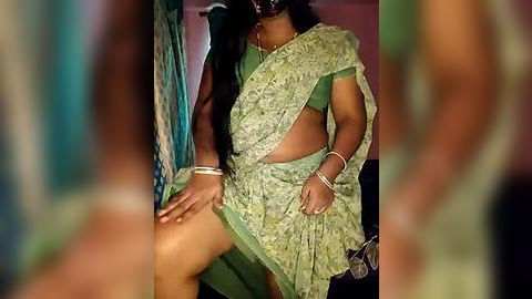 Media: Video of a woman in a green saree with intricate patterns, lifting the saree to reveal her thigh. She wears a black mask, bracelets, and a necklace. Background is blurred.