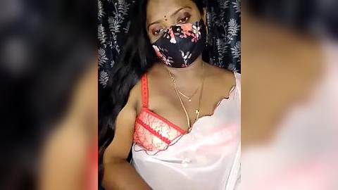 Media: Video of a South Asian woman with long black hair, wearing a black floral mask, red lace bra, and white blouse, with a floral backdrop.