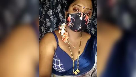 Media: Video of a South Asian woman with medium brown skin and dark hair, wearing a blue saree, floral mask, and intricate jewelry, against a dark floral-patterned backdrop.