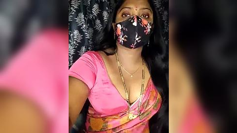 Media: Video of a South Asian woman with medium skin tone, wearing a pink saree, floral face mask, and gold necklace. Background features dark floral curtains.