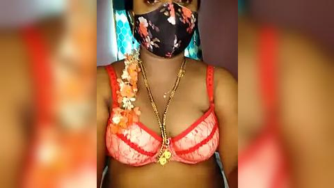 Media: Video of a dark-skinned woman wearing a floral pink bra, black mask, and gold necklace, set against a blurred teal and orange background.