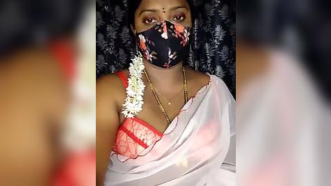 Media: Video of a South Asian woman with medium brown skin, wearing a red lace bra, a floral mask, and a white saree with a gold necklace. Background shows dark patterned fabric.