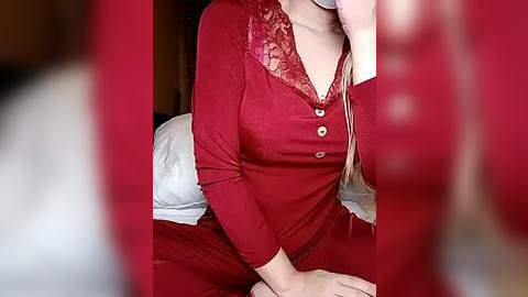 Media: Video of a woman in a red, long-sleeved nightgown with lace details, sitting on a bed with white sheets, partially blurred in the foreground.