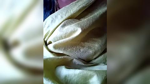 Media: A close-up video of a person's face partially covered by crumpled, beige fabric, possibly a blanket or clothing. The fabric's texture is soft and slightly wrinkled. The background is blurred, focusing attention on the fabric and the person's features.