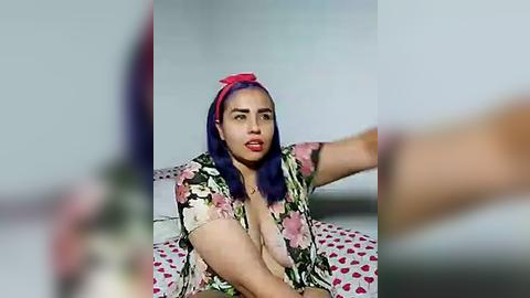 Media: Video of a dark-skinned woman with purple hair and a red headband, wearing a low-cut floral dress, sitting on a bed with red heart-patterned sheets. The background is blurred, with a white wall.