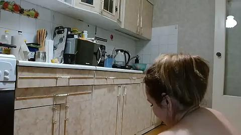 Media: Video of a woman with brown hair tied in a bun, wearing a yellow apron, cooking in a beige kitchen with white cabinets and tiled backsplash.