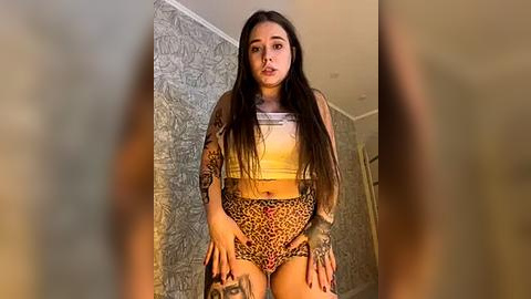 Media: Video of a young woman with long, dark hair, wearing a leopard-print crop top and shorts, standing in a room with gray-patterned wallpaper.