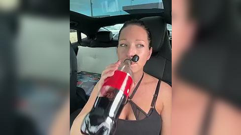 Media: Video of a woman with dark hair, wearing a black tank top, drinking from a red and black water bottle while seated in a car.