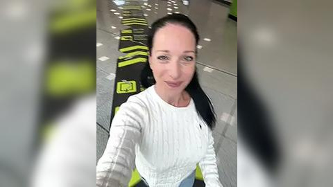 Media: A video of a smiling woman with long black hair, wearing a white knit sweater, standing in a modern, light-filled space with black and green patterns on the floor.