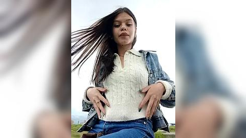 Media: A video of a young woman with long black hair, wearing a white knit top and blue jeans, sitting on grass outdoors, with blurred figures in the background.