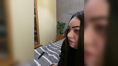 Media: Video of a young woman with long dark hair, wearing a black top, sitting on a bed with a gray and white striped duvet. Background includes a wooden wardrobe and a potted plant on a shelf.