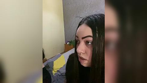 Media: Video of a young woman with long black hair, dark eyebrows, and a septum piercing, partially obscured by a blurred figure, in a dimly lit, cluttered bedroom with a gray wall and a yellow blanket.