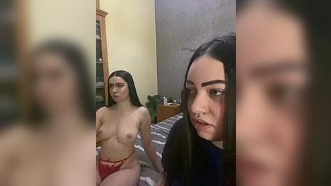 Media: Video of two young women in a dimly lit room: one topless with red lace panties, another with long black hair, a nose ring, and a neutral expression.