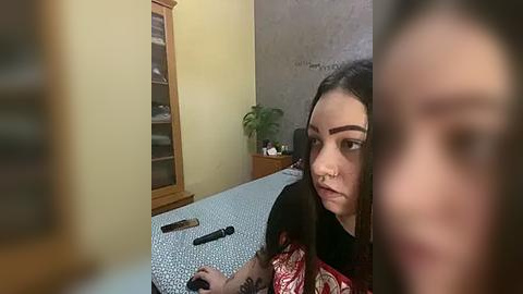 Media: Video of a young woman with long dark hair, wearing a red shirt, looking contemplative in a cluttered room with a wooden cabinet, plant, and a bed.