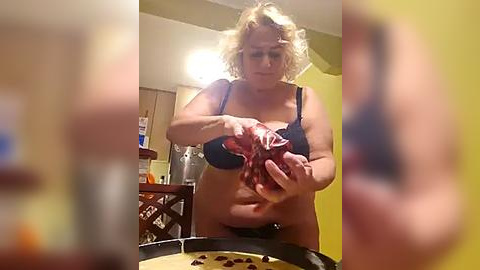 Media: Video of a plus-sized woman with curly blonde hair, wearing a dark bra, holding a red object (possibly meat) over a wooden table, in a dimly lit, cluttered kitchen with yellow walls.