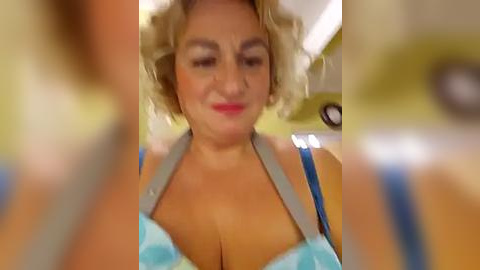 Media: A blurry video shows a middle-aged woman with short curly blonde hair, wearing a light blue bra with polka dots, and a grey apron. The background is indistinct.