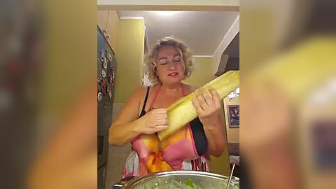 Media: Video of a middle-aged, fair-skinned woman with curly blonde hair, wearing a pink apron, cutting a large yellow squash in a well-lit kitchen.