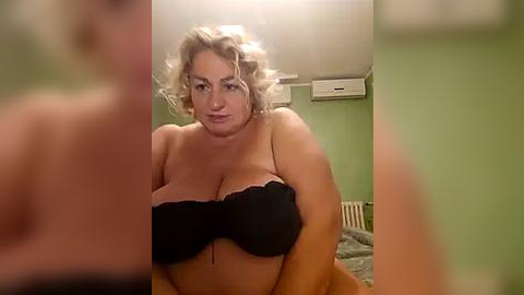 Media: Video of a plus-sized woman with blonde curly hair, wearing a black strapless bra, standing in a room with green walls and an air conditioner.