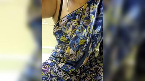 Media: A video of a woman wearing a colorful, sleeveless dress with a bold, tropical leaf pattern in blue, yellow, and white. Her skin tone is light, and she has a black bra strap visible. The background is out of focus, showing a light yellow wall.