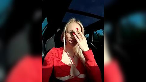 Media: Video of a blonde woman in a red top, sitting in a car, adjusting her glasses, with a blurred background and red-tinted edges.