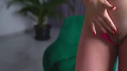 Media: A video showing a close-up of a fair-skinned woman's hand with red-painted nails, touching her shaved vulva. The background features a blurred green chair and a potted plant.