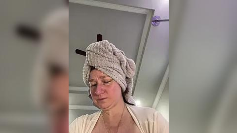 Media: A video of an older woman with light skin, wearing a beige robe and a fluffy, gray towel turban with two black antlers on top. She has a solemn expression, and the background shows a modern, gray-toned bathroom with a white ceiling.