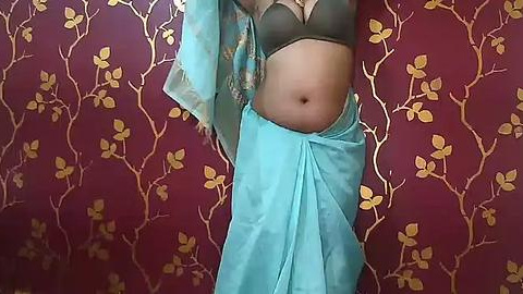 Media: Video of a woman's midsection, wearing a turquoise sari with a dark green bra, against a red wallpaper with gold floral designs. Her belly is visible, emphasizing a slender, toned physique.