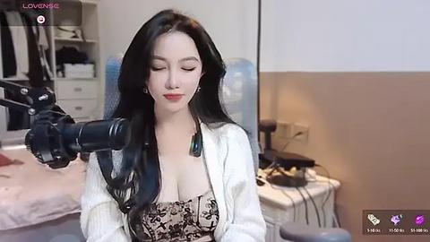 Media: Video of a fair-skinned woman with long black hair, wearing a low-cut black top, sitting in a well-lit room with a white robe and a large green necklace.