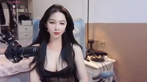 Media: A video of an East Asian woman with long black hair and fair skin, wearing a black top, using a professional camera to film herself in a bedroom with a bed, desk, and blue chair.