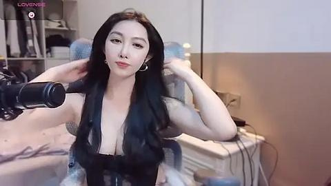 Media: Video of a young East Asian woman with long black hair, wearing a black, low-cut dress, recording herself with a camera in a cluttered, beige-walled bedroom.