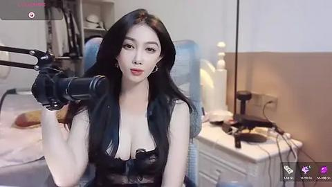 Media: A video of an East Asian woman with long black hair, wearing a black lace bra, sitting in a bedroom with a white bed, water bottles, and a lamp.