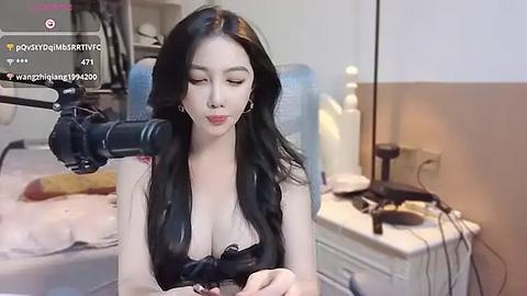 Media: Video of a young Asian woman with long black hair, wearing a black top, using a professional camera on a tripod in a modern bedroom with beige walls, a bed, and a desk with electronics.