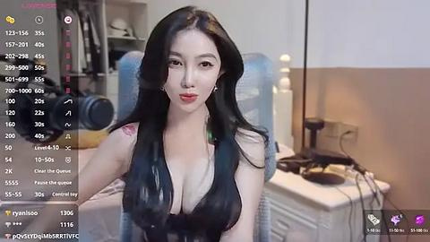 Media: Video of an East Asian woman with long black hair, fair skin, wearing a revealing black dress, sitting in a bedroom with a bed, nightstand, and water bottles.