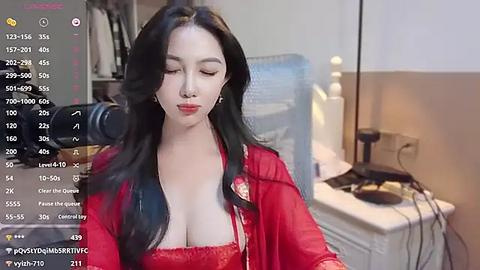 Media: Video of a young East Asian woman with long black hair, fair skin, and red lipstick, wearing a red lace top, standing in a cluttered room with a white chair, a lamp, and a camera on a tripod.