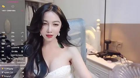 Media: Video of a young East Asian woman with long black hair, wearing a strapless white dress, standing in a modern bedroom with white furniture and a desk.