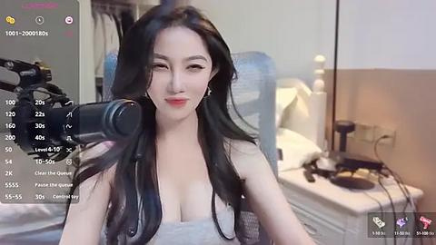 Media: Video of an Asian woman with long black hair, fair skin, and large breasts, wearing a low-cut gray top, speaking into a microphone in a bedroom with white walls and a bed.