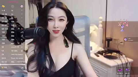 Media: Video of an Asian woman with long black hair and fair skin, wearing a black sheer top, sitting in front of a camera in a bedroom.