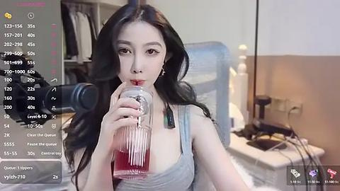 Media: Video of an East Asian woman with long black hair, wearing a gray tank top, drinking from a glass jar, in a dimly lit bedroom with a bed, chair, and desk.