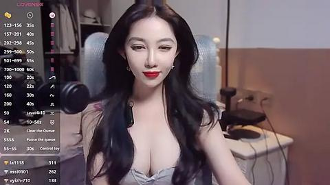 Media: Video of a smiling East Asian woman with long black hair, red lipstick, wearing a revealing, low-cut, light gray top, sitting in a dimly-lit room with a chair and desk in the background.