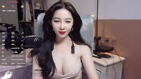 Media: Video of an East Asian woman with long, wavy black hair, fair skin, and red lipstick, wearing a low-cut beige top, sitting in a dimly lit room with a camera focused on her.