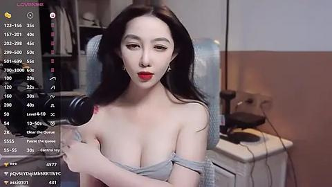 Media: Video of a fair-skinned, East Asian woman with long black hair and red lipstick, wearing a gray off-shoulder top, sitting in front of a microphone, with a cluttered room background.