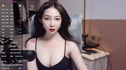 Media: Video of an East Asian woman with long black hair, fair skin, and red lipstick, wearing a black tank top, sitting in a dimly lit room with a camera in front of her, streaming on Twitch.