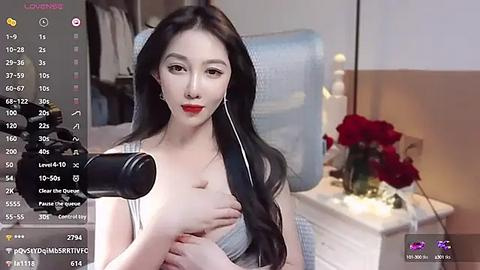 Media: Video of an East Asian woman with long black hair, wearing a white tank top, holding a microphone, in a bedroom with a bed, a table with red roses, and a blue chair.