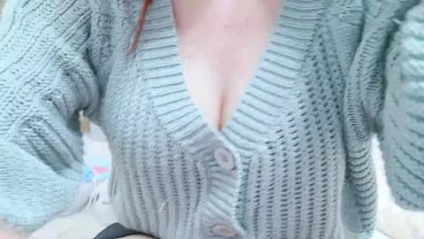 Media: Video of a light-skinned woman with long red hair, wearing a light grey, knitted cardigan with a deep V-neck, showcasing ample cleavage.