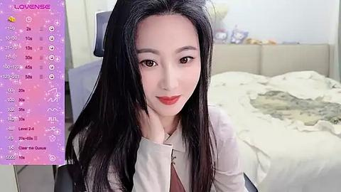 Media: A video of a young Asian woman with long black hair, fair skin, and red lipstick, sitting on a bed with a messy white blanket. The background shows a room with a gray headboard and a pink \"LOVENSE\" screen.