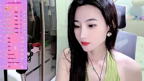 Media: A video of an East Asian woman with long black hair, wearing a sleeveless green top and red lipstick, sitting indoors, with a background of a bedroom with a closet and a bed.