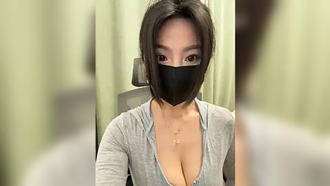 Media: Video of a young Asian woman with shoulder-length black hair, wearing a black face mask, light grey V-neck top, and a gold necklace, sitting in a dimly-lit room with light green curtains.