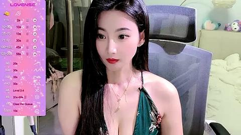 Media: Video of a young East Asian woman with long black hair, wearing a green floral halter top, sitting in a gray office chair. Background features a cluttered room with a purple wall and a bed.