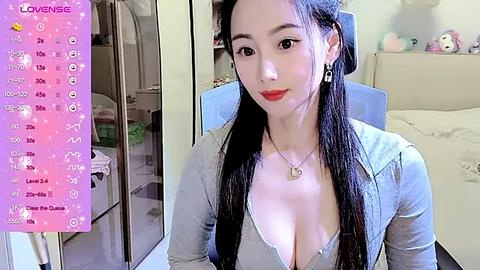 Media: Video of an East Asian woman with long black hair, wearing a low-cut denim shirt revealing cleavage, sitting in a bedroom with a bed and pastel decor.